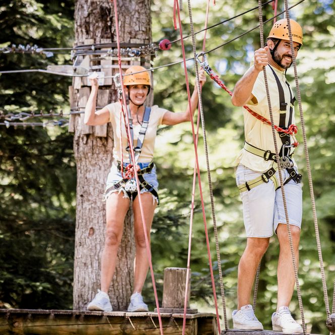 Eine Frau und ein Mann mit orangefarbenen Helmen und Klettergurten stehen gesichert auf Plattformen zwischen Seilen im Hochseilgarten, umgeben von Nadelbäumen