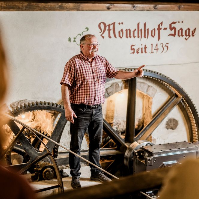 Ein Mann im karierten Hemd steht erklärend vor einer weißen Wand mit der Aufschrift Mönchhof-Säge seit 1435 und einem großen Zahnrad, im Vordergrund Zuhörer