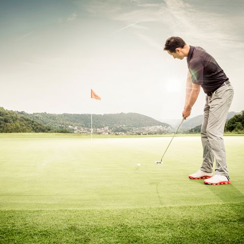 Ein Golfer beim Abschlag auf dem Golfplatz. Im Hintergrund die Fahne am Flaggenstock zur Kennzeichnung des Lochs für den Spieler.
