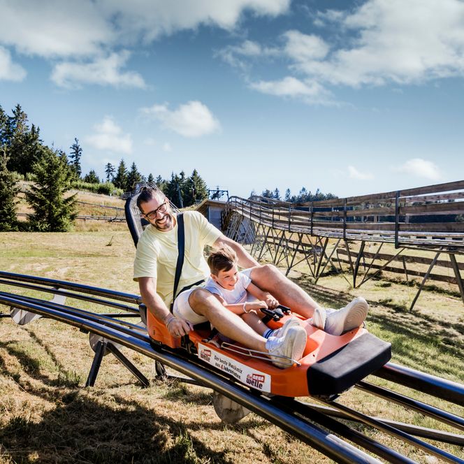 Ein Mann im gelben Shirt und ein Junge fahren lachend in einem orangefarbenen Coaster-Wagen auf einer kurvigen Schienenbahn über eine Wiese, dahinter Wald