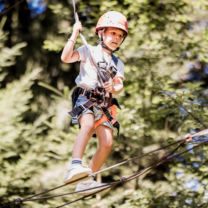 Ein Junge mit orangefarbenem Helm, Klettergurt und weißem Shirt balanciert konzentriert auf einem Drahtseil und hält sich an einem Sicherungsseil fest, dahinter Wald