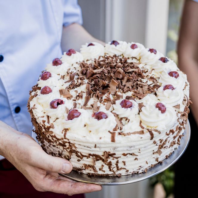 Nahaufnahme einer runden Schwarzwälder Kirschtorte mit Sahnetupfen, roten Kirschen und Schokoladenraspeln, gehalten auf einer Platte von einem Mann im hellen Hemd