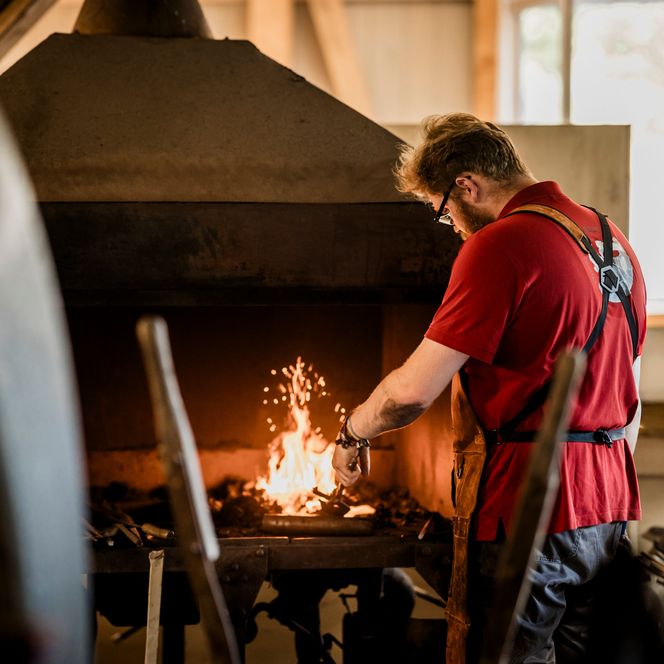 Ein Schmied mit Lederschürze steht an der Esse, aus der Flammen und Funken schlagen, und erhitzt ein Werkstück im Feuer