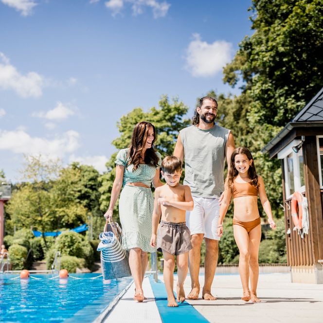 Ein Mann, eine Frau und zwei Kinder in Badekleidung gehen am Rand eines Schwimmbeckens, links eine blaue Wasserrutsche, im Hintergrund Bäume