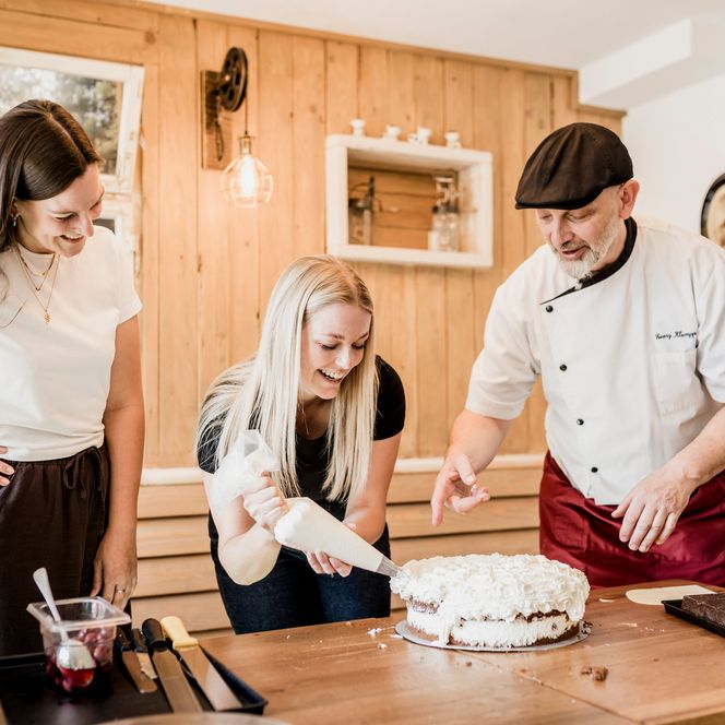 Eine blonde junge Frau spritzt mit einem Spritzbeutel Sahne auf eine Torte, ein Konditor mit Mütze zeigt darauf, links schaut eine weitere Frau lächelnd zu