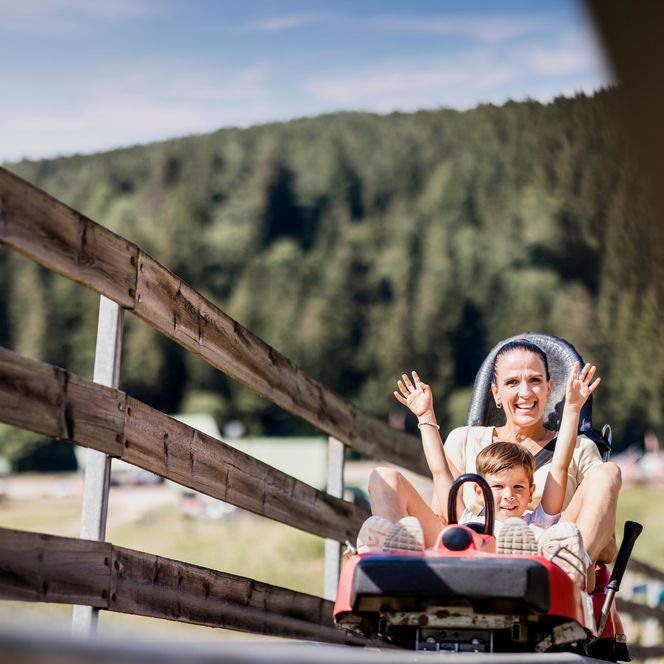 Eine Frau hebt lachend beide Arme, vor ihr sitzt ein Junge am Steuerhebel eines Coaster-Wagens, im Vordergrund ein Holzgeländer, dahinter bewaldeter Hang
