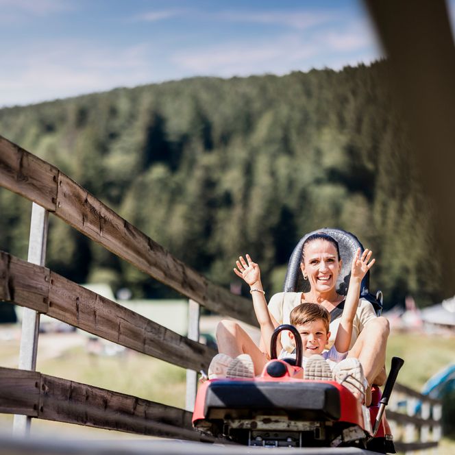 Eine Frau hebt lachend beide Arme, vor ihr sitzt ein Junge am Steuerhebel eines Coaster-Wagens, im Vordergrund ein Holzgeländer, dahinter bewaldeter Hang