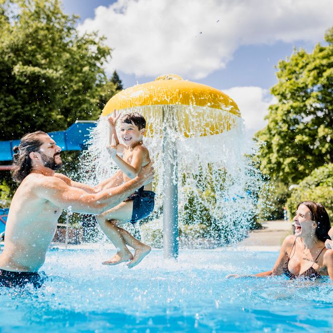 Ein Mann hebt einen jubelnden Jungen aus dem Wasser hoch, rechts lachen eine Frau und ein Mädchen, im Hintergrund die gelbe Pilzdusche mit Wasservorhang