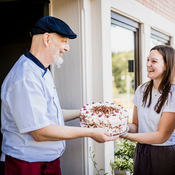 Ein Konditor mit dunkler Mütze reicht zwei lachenden jungen Frauen vor einem Café eine fertige Schwarzwälder Kirschtorte mit Sahne, Kirschen und Schokoraspeln
