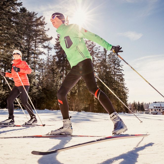 Aus niedriger Perspektive laufen ein Mann in grüner Jacke und eine Frau in oranger Jacke auf einer Loipe, die Sonne strahlt über verschneiten Bäumen und einem Schwarzwaldhaus