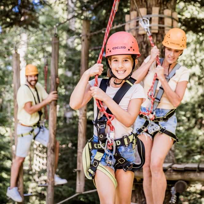 Ein lachendes Mädchen mit rosa Helm und Klettergurt hält sich an einem roten Seil fest, dahinter eine Frau und ein Mann mit orangefarbenen Helmen zwischen Bäumen
