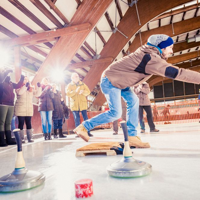 Ein Mann mit Mütze wirft in gebückter Haltung einen Eisstock über die Eisbahn, am Rand jubelt eine Gruppe Zuschauer in Winterkleidung